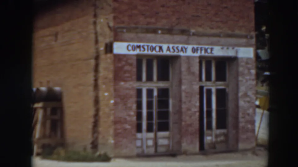 1937 Virginia City home-movie frame showing the Comstock Assay Office sign above a weathered brick facade with double doors and windows.