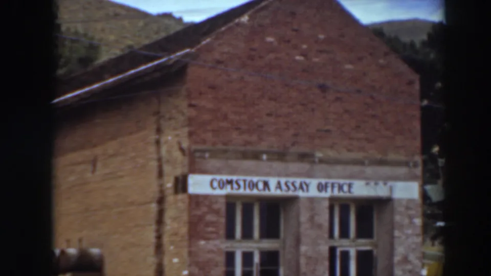 Near the end of the pan, the frame includes the Comstock Assay Office sign, upper brick gable, roofline, and hills behind the building.