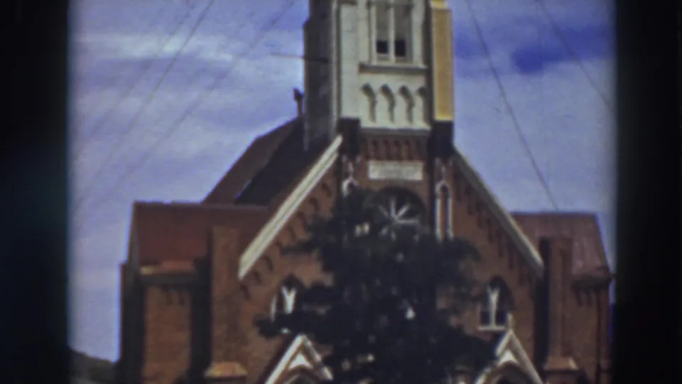 Late-frame 1937 view tilting upward on the Virginia City cathedral, showing the steeple section above the brick facade and tree.