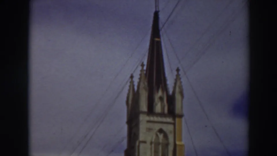 1937 8mm view of an ornate building in Virginia City, California, with a pale yellow car parked in front and overhead wires crossing the sky.