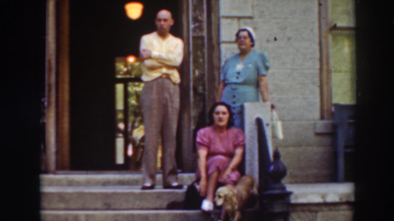 1937 home-movie frame in Virginia City, California, showing two adults standing on a porch and a woman seated beside a small dog.