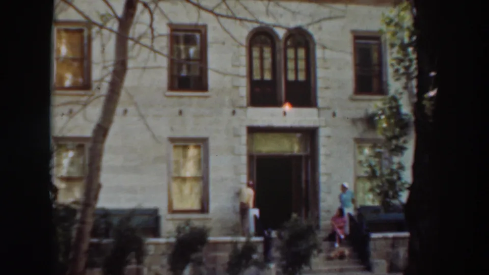 Wide 1937 8mm view of three adults and a small dog near the entrance of a large Virginia City, California building.