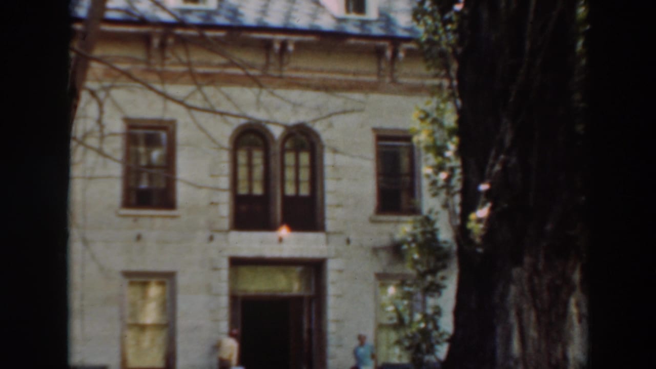 Tan-yellow two-story house partly framed by dark tree trunks, with people near the entrance.