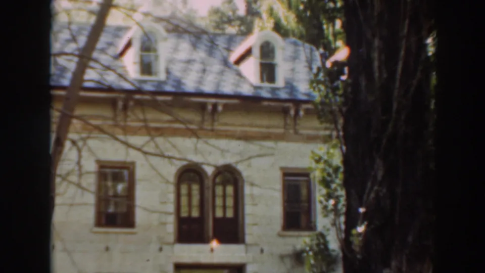 The camera holds on the house facade, showing arched upper windows, a dark roof with dormers, bare branches, and a large tree trunk in front.