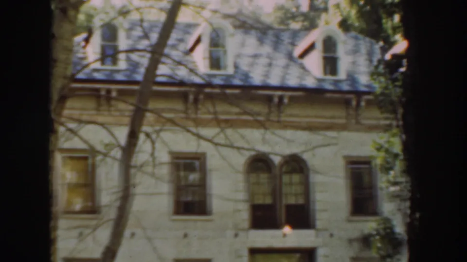 A closer 1937 home-movie frame shows the tan-yellow facade, paired arched windows, rectangular windows, roofline, and trees crossing the view.