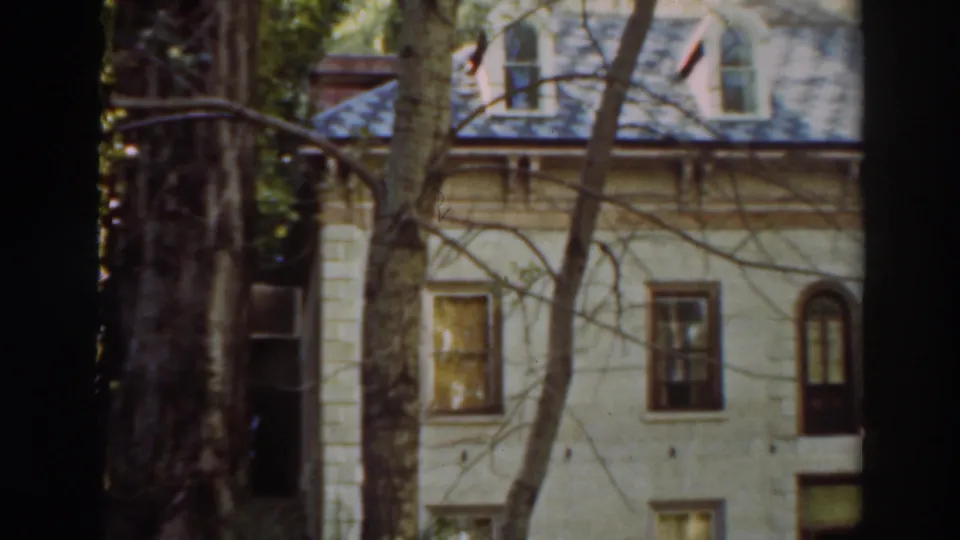 1937 8mm frame of a pale house in Virginia City, California, seen through trees with dormer windows and a dark roofline.