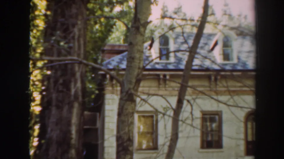 The camera holds on the Virginia City house exterior, with tree trunks and branches partially blocking the pale facade and roof.