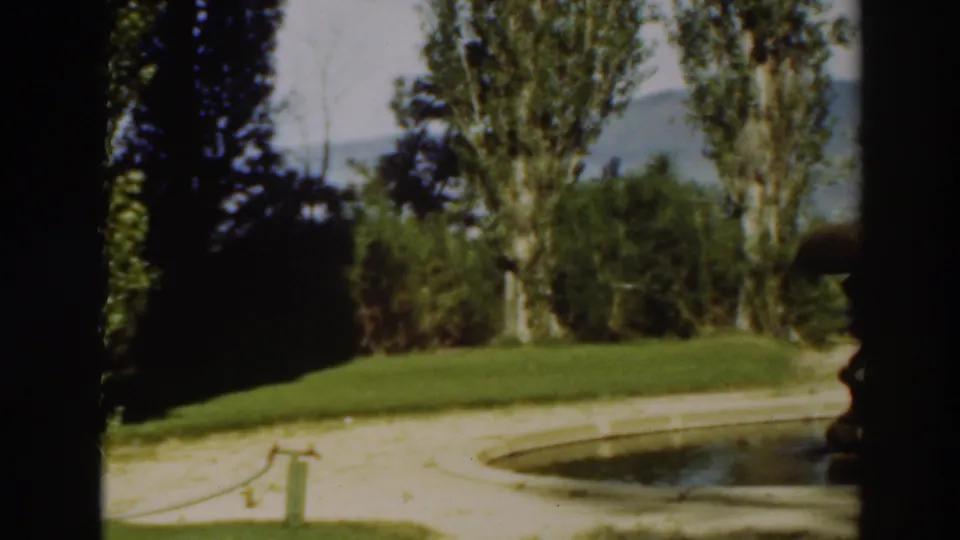 The scene shifts to landscaped grounds in Virginia City, California, with green lawn, garden path, fountain edge, tall trees, and distant hills.