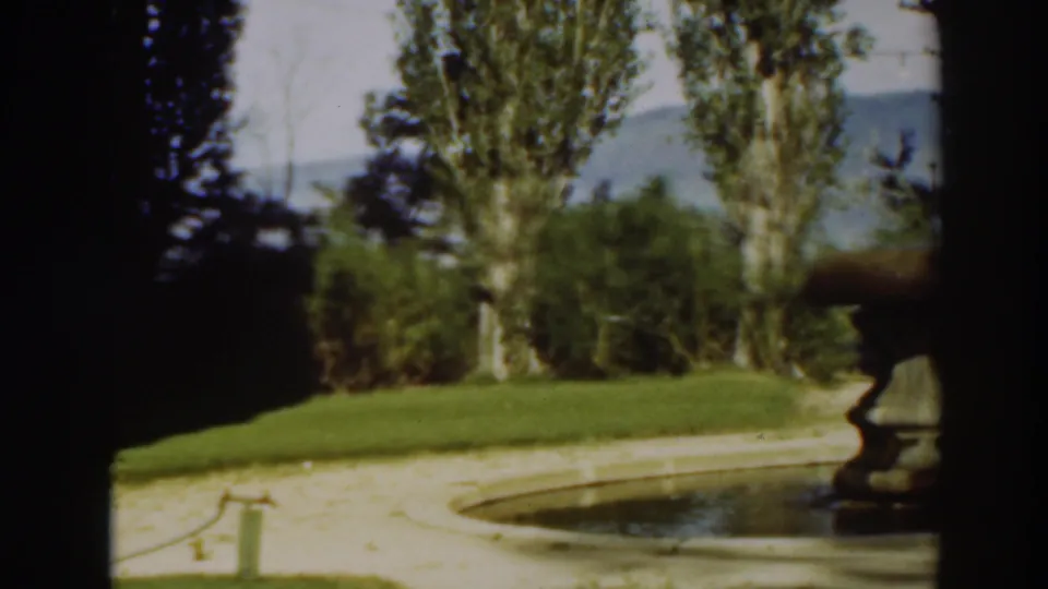 1937 8mm frame from Virginia City, California, showing a circular stone fountain near grass, tall trees, and a distant hillside.