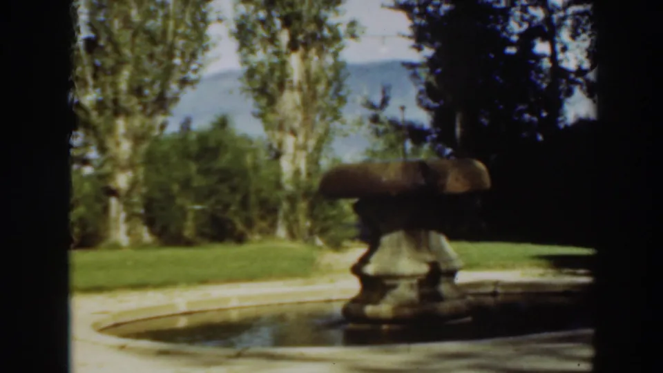 1937 8mm frame from the garden scene, showing the stone fountain, basin, bright grass, tall trees, and dark foliage.