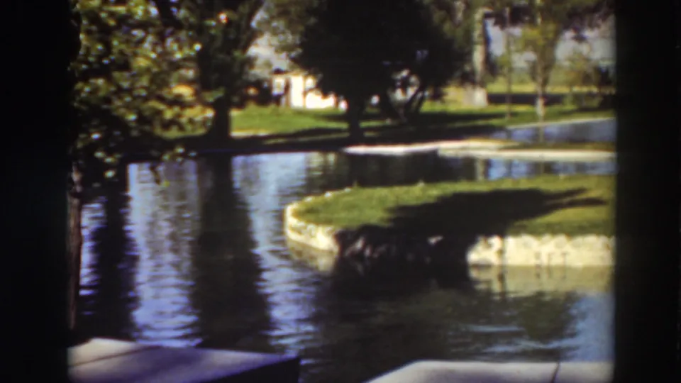 Landscaped water feature with rippled reflections, a curved grass edge, trees, and bright lawn areas in vintage 8mm footage.