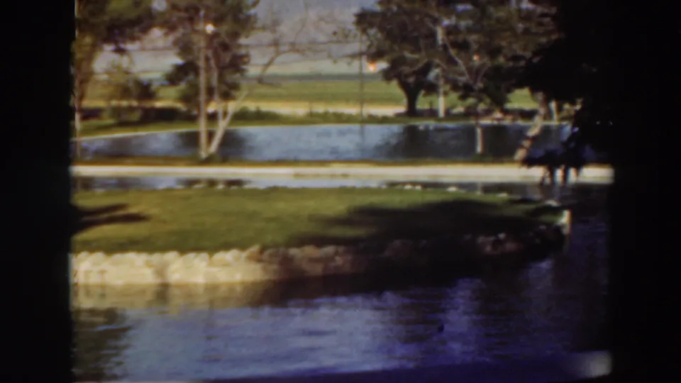 1937 home-movie view of a grassy waterside park in Virginia City, California, with a possible distant person near the trees.