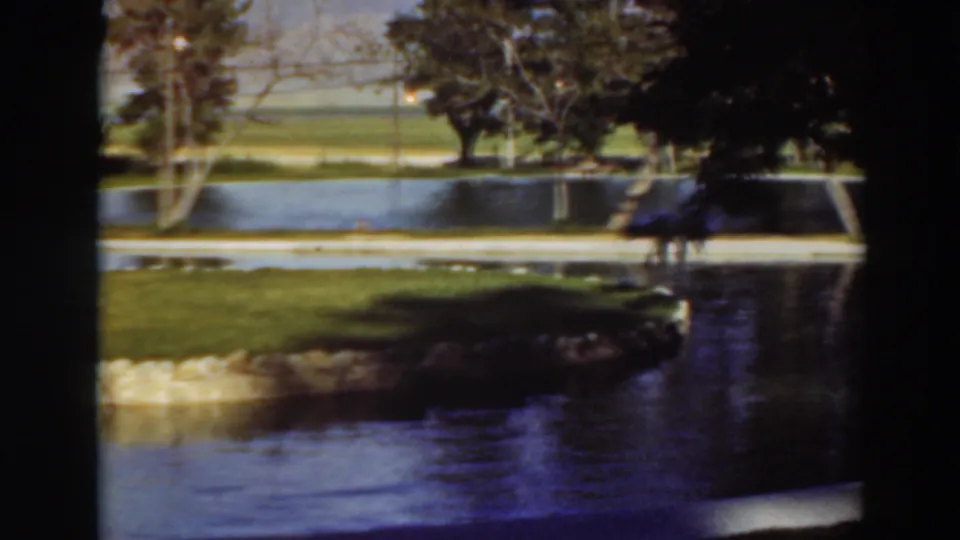 A soft 8mm frame from the 1937 Virginia City, California clip shows water, lawn, trees, and a possible distant person.