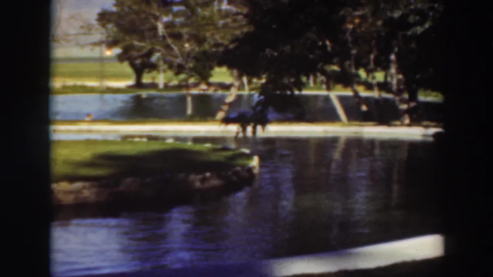 Home-movie frame of the supplied 1937 Virginia City, California park pond scene with trees, reflected water, and distant people.