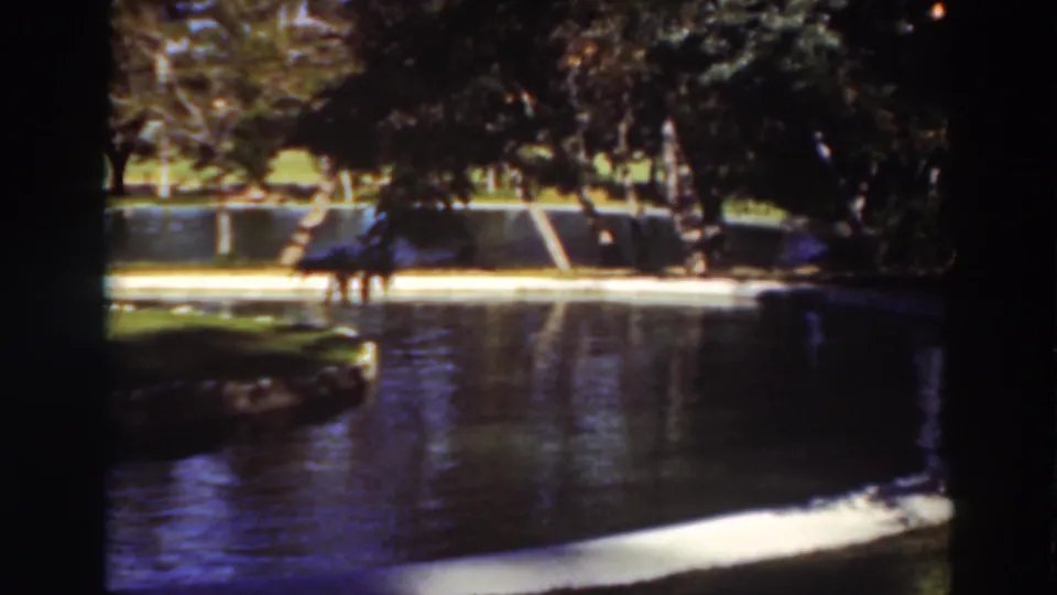 Faded 8mm frame from the supplied 1937 park scene, showing pond water, a grassy island, mature trees, and distant visitors.