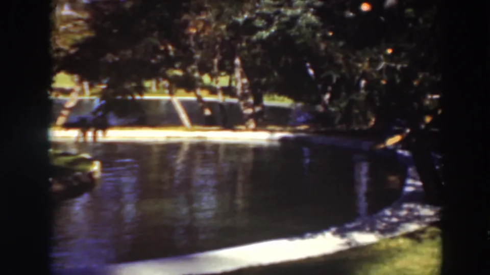 Late frame of the supplied 1937 Virginia City, California park pond shot, with heavy shade, bright reflections, and small figures across the water.
