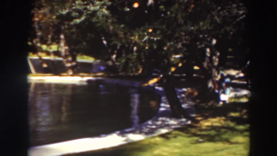 1937 8mm park pond scene in Virginia City, California, with shaded trees, a concrete border, grass, and a small person near the right edge.