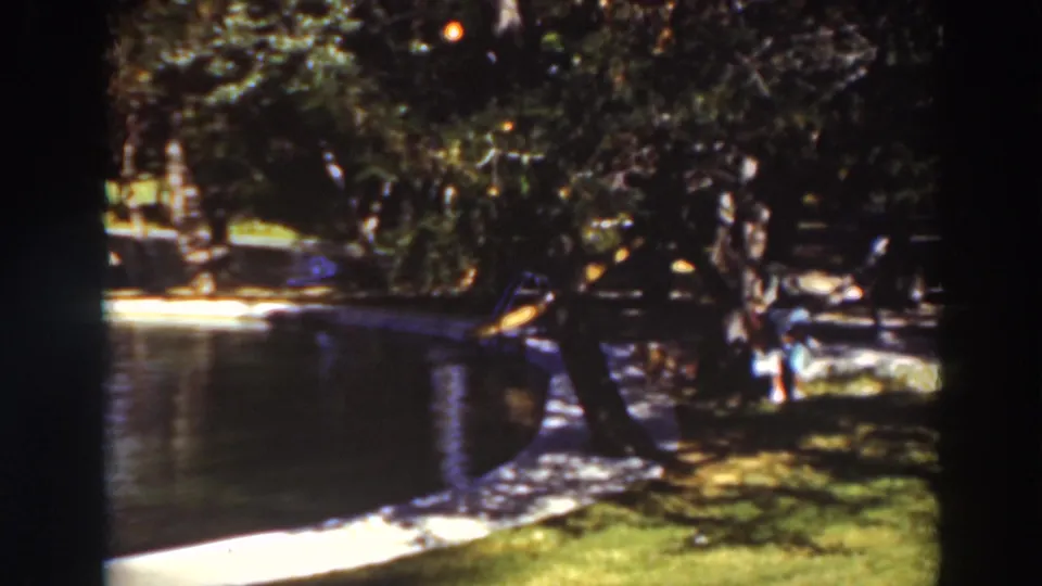 Virginia City, California park pond in 1937, with soft 8mm color, a grassy bank, trees, and a distant small figure by the water.