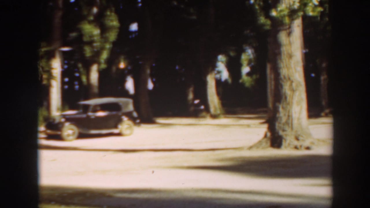 1937 8mm frame showing a dark parked car with a pale roof beside tall trees in Virginia City, California.
