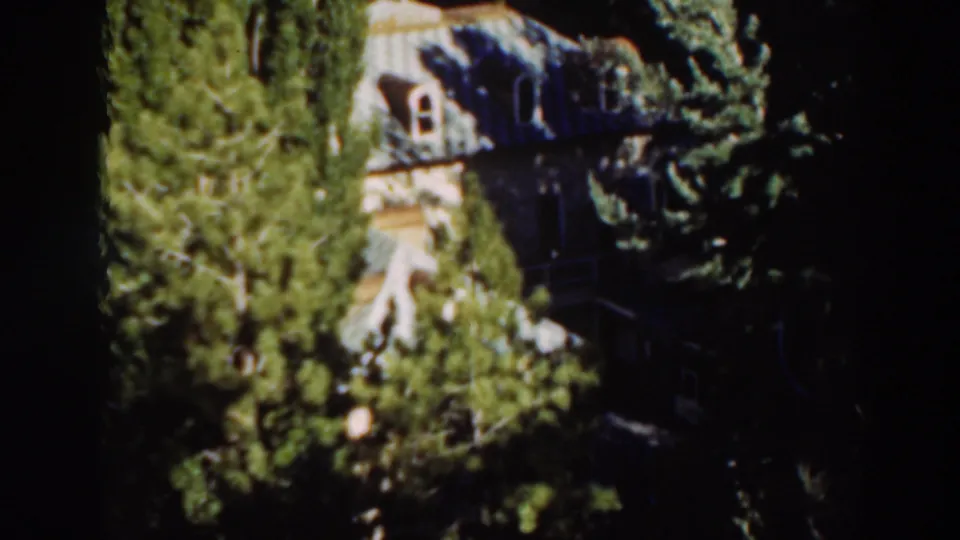 A pale large house appears partly obscured by trees in this 1937 Virginia City home-movie frame, with windows visible through foliage.