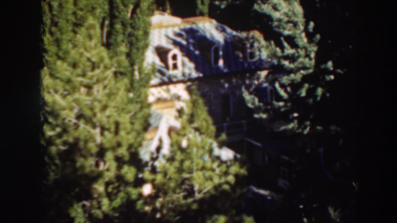 1937 8mm frame from Virginia City, California showing a pale house with arched windows partly obscured by evergreens.