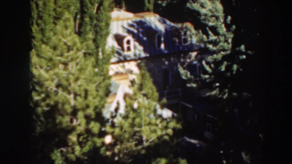 1937 8mm frame from Virginia City, California showing a pale house with arched windows partly obscured by evergreens.