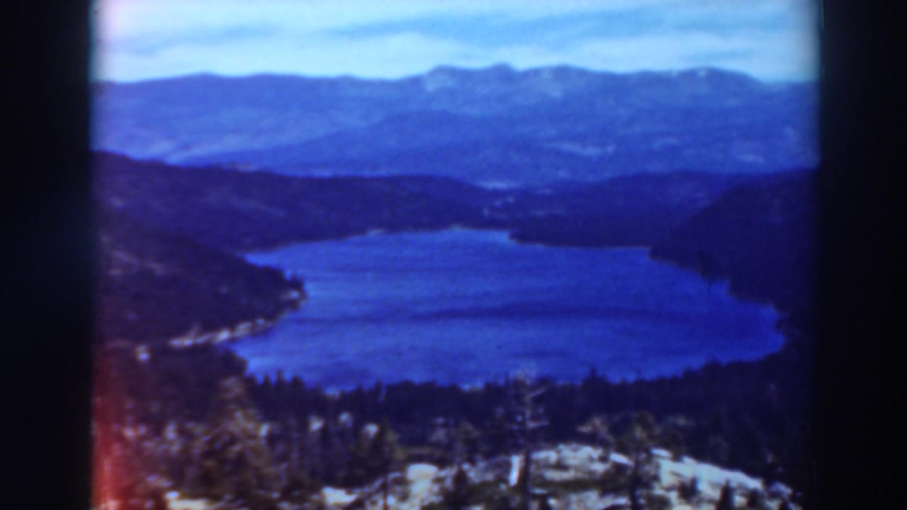 Blue lake below forested mountain slopes with distant ridges and pale sky.