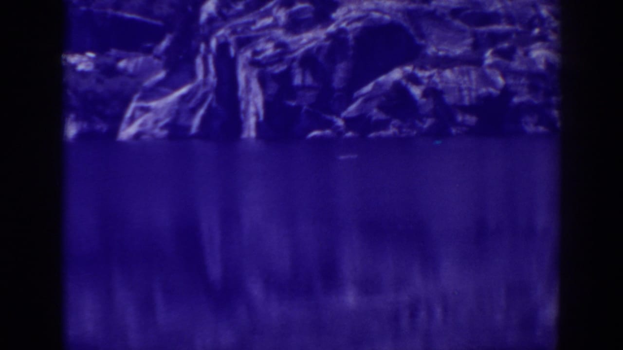 Dim blue view of still lake water below a rocky tree-lined shore.