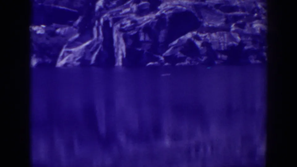 Blue-shifted 1938 8mm view at Angora Lakes showing calm water, rocks, and conifers along the shoreline.