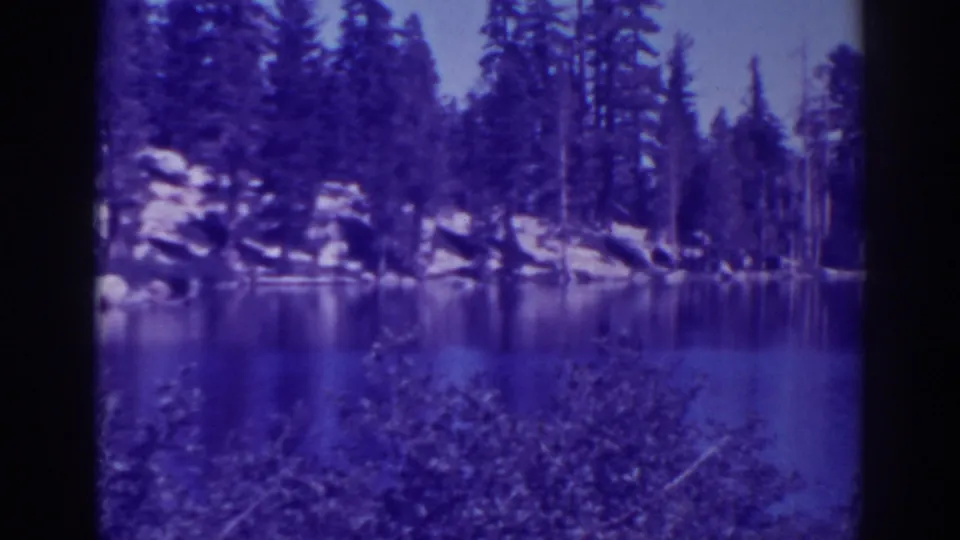 1938 home-movie frame of Angora Lakes with foreground brush, reflective water, boulders, and dense pine trees.