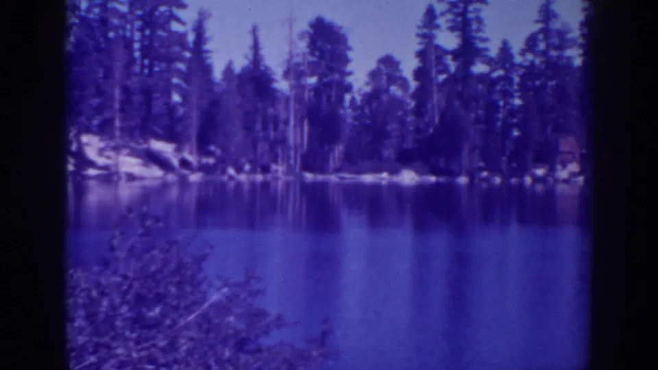 1938 8mm frame of Angora Lakes showing foreground brush, still water, and tall pines along the opposite shore.