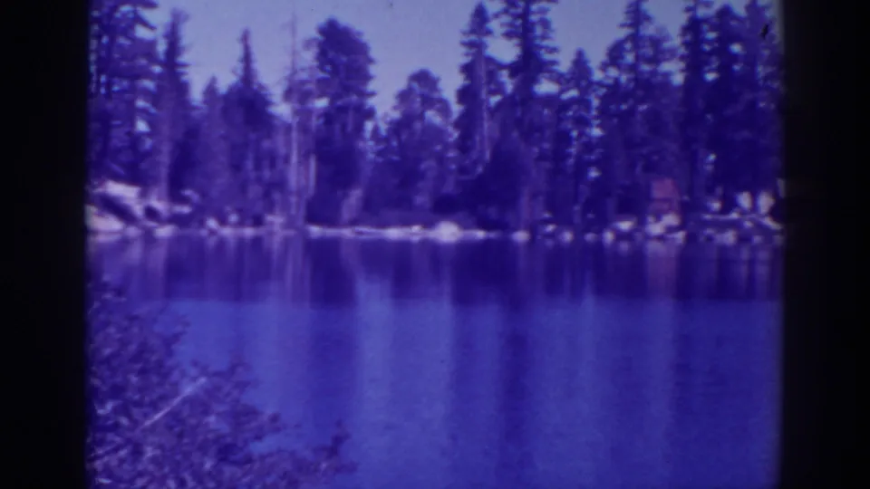 Vintage 8mm view of Angora Lakes with a calm reflective surface, pine trees, shoreline rocks, and strong purple-blue cast.