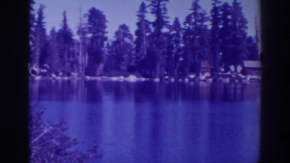 1938 Angora Lakes view across calm water toward pine trees and small cabins, seen in soft purple-blue 8mm footage.