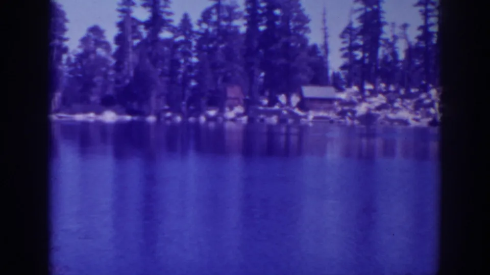 1938 Angora Lakes footage showing a wide water view toward a wooded shoreline with rocks and distant cabins.