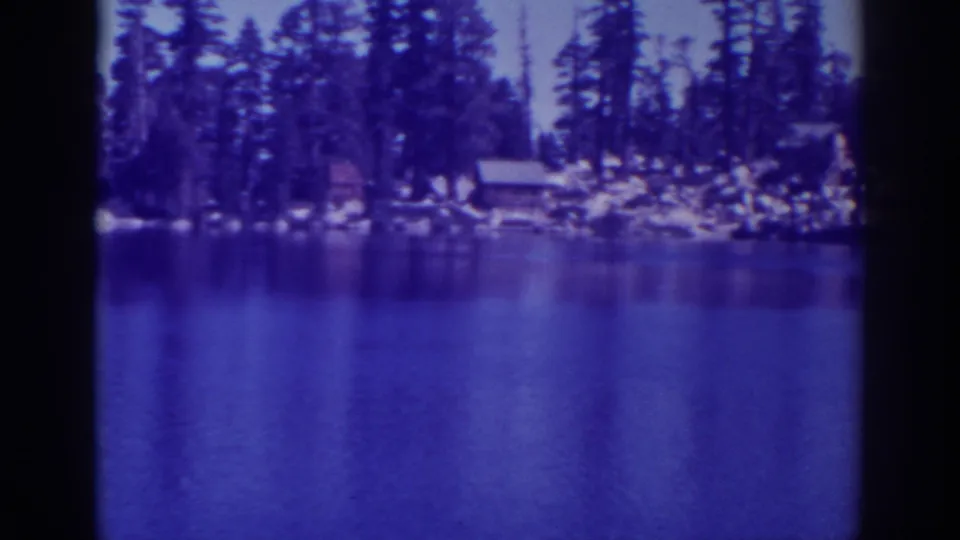 1938 Angora Lakes scene with calm foreground water, pine-covered shore, small cabins, and rocky edges.