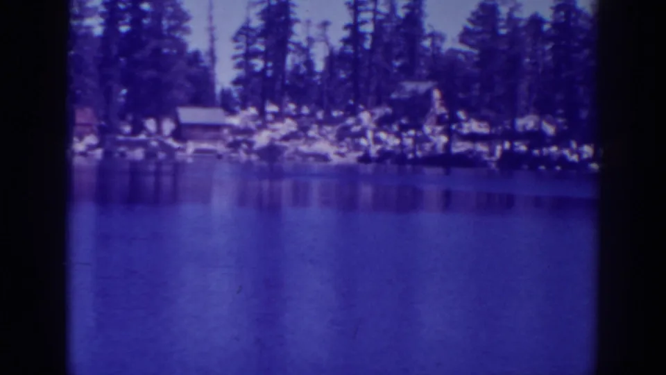 Angora Lakes shoreline seen across calm water, with pine forest, rocks and small cabins in soft 8mm color.