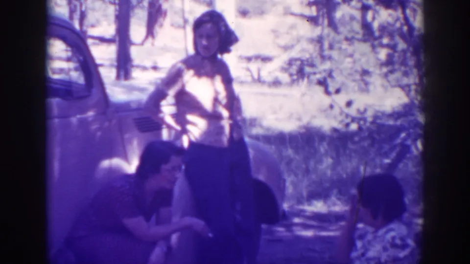 At Angora Lakes, three women appear near a parked vintage car in a sunlit wooded area, with one standing and two seated.
