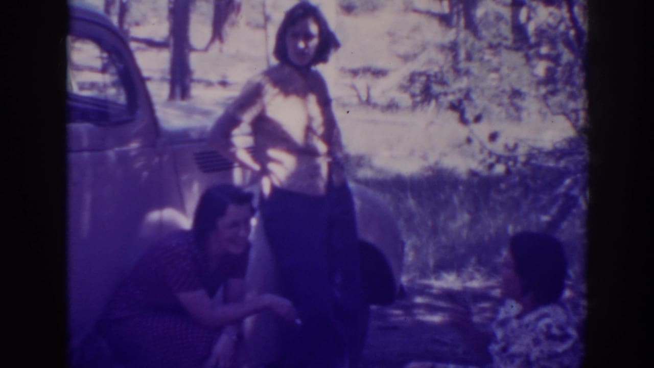 Three women near a classic car in a wooded outdoor setting.