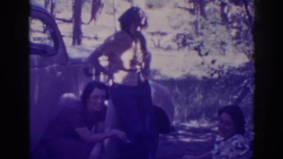 A woman stands with hands on hips beside a classic car while two women sit on the ground at Angora Lakes in 1938.