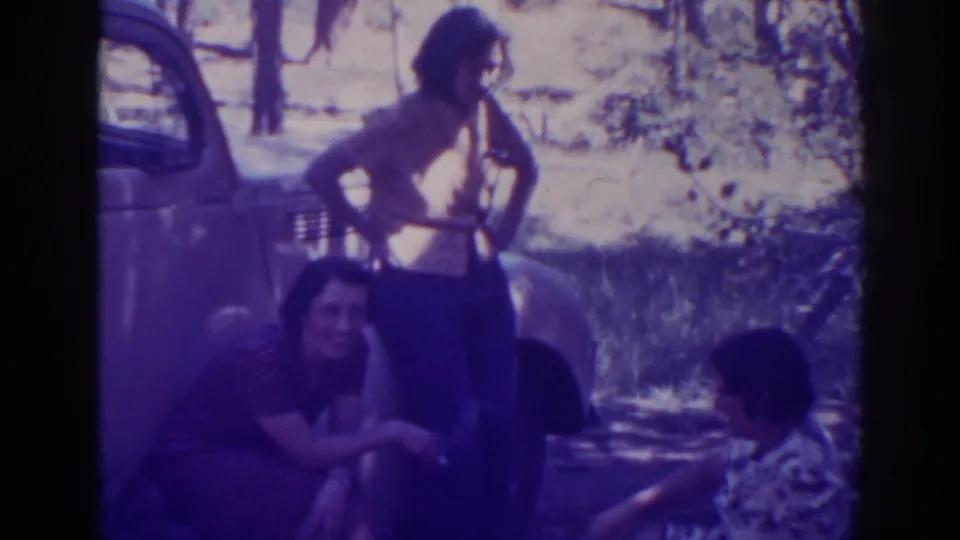 Three women remain grouped beside the classic car, with bright forest light and strong purple color cast typical of aged 8mm film.