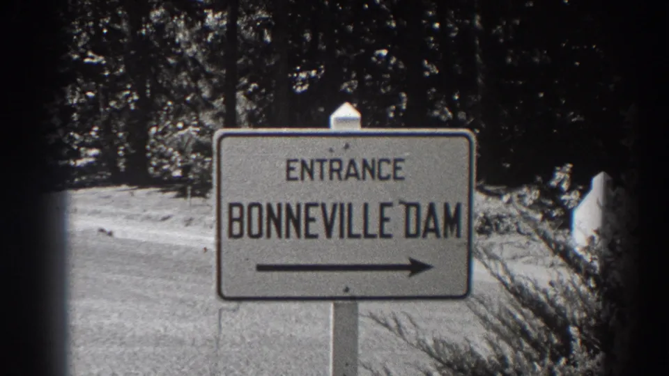 Final frame holds on the 1938 Bonneville Dam entrance sign in Oregon, with forest and roadside brush around it.