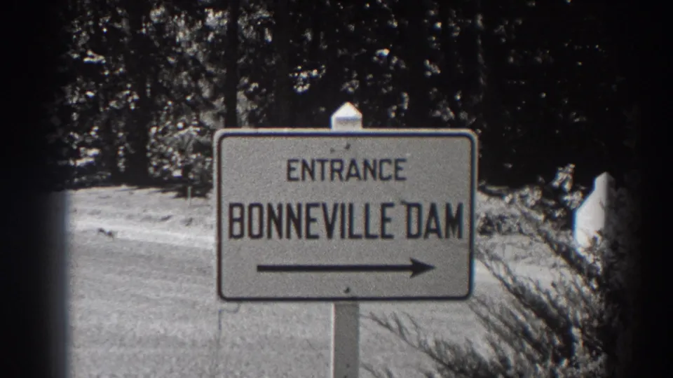 1938 8mm frame at Bonneville Dam showing a white entrance sign with a right-pointing arrow along a wooded roadside.