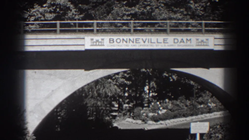 1938 home-movie frame showing the Bonneville Dam sign across a bridge, with a large concrete arch and landscaped trees below.
