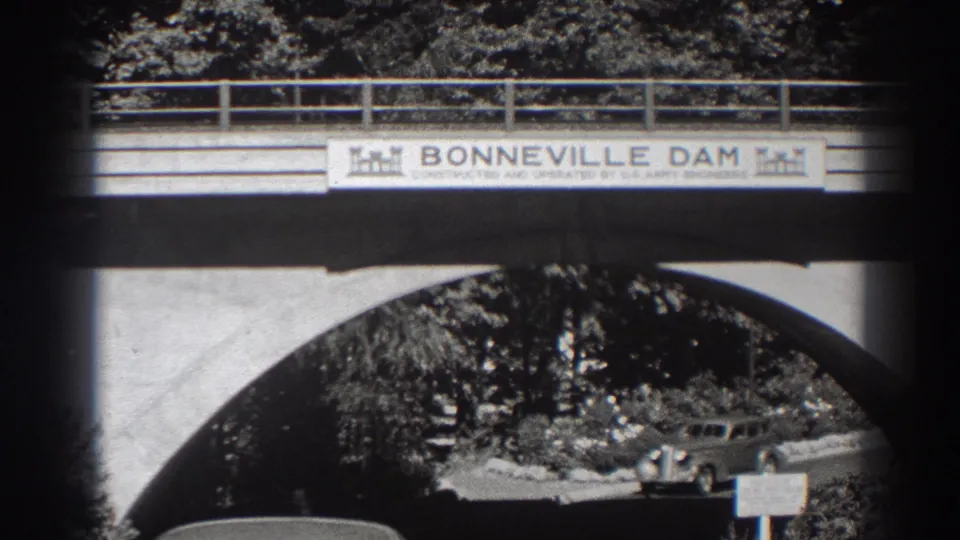 A vintage car moves under the Bonneville Dam bridge arch in Oregon, with the dam sign, railing, trees, and road visible.