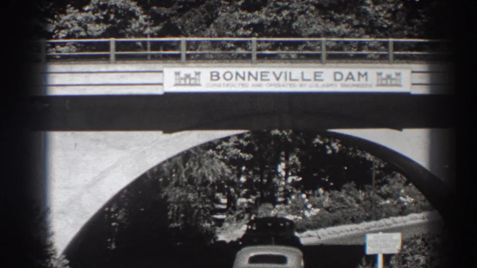 The car continues beneath the Bonneville Dam bridge arch, framed by the sign, concrete span, railing, trees, and roadside marker.