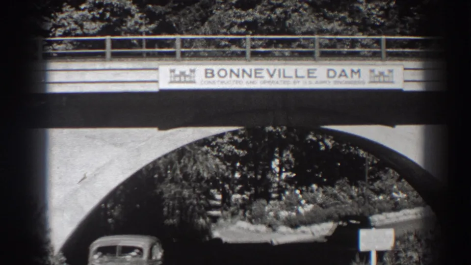 At Bonneville Dam, a rounded 1930s automobile moves under the signed bridge, with trees and a roadside sign in the background.