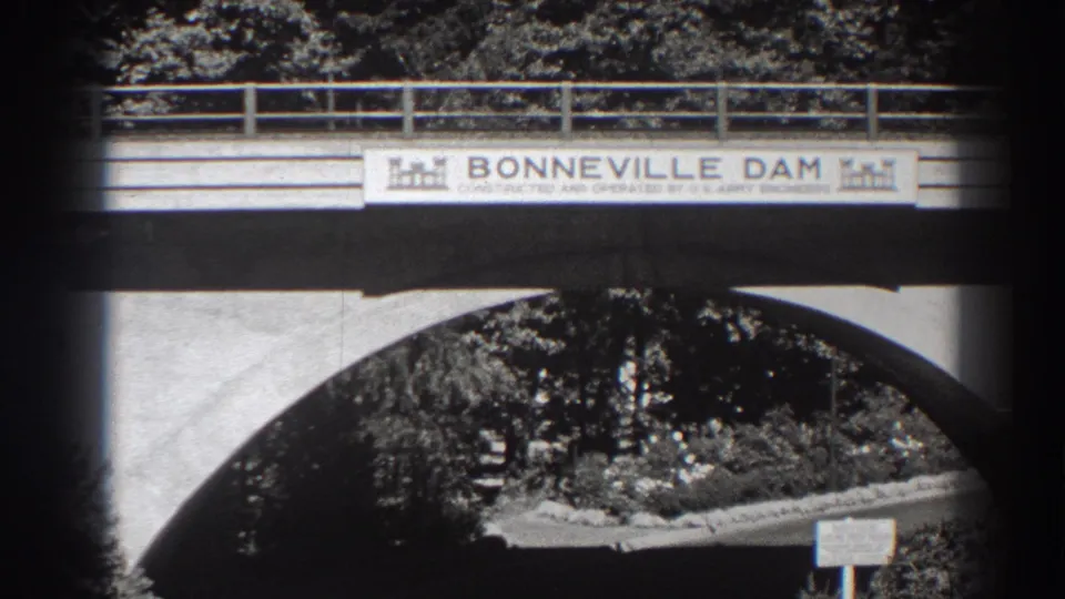 Black-and-white 8mm frame of the Bonneville Dam bridge sign over a concrete arch, with the roadway mostly clear below.