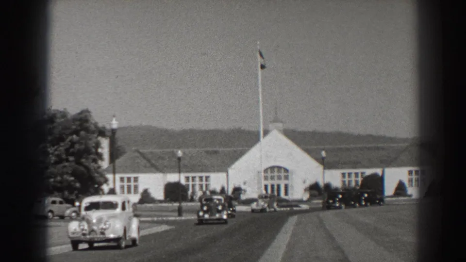 Wide 1938 Bonneville Dam exterior view showing a white public building, central flagpole, roadway, and period automobiles.