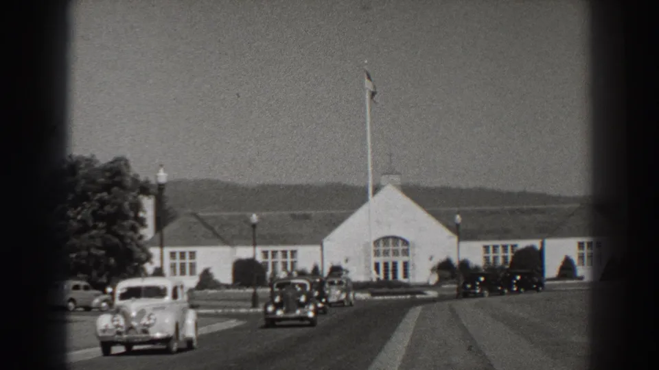 Bonneville Dam area road scene with a white building, flagpole, distant hills, and 1930s automobiles moving or parked nearby.
