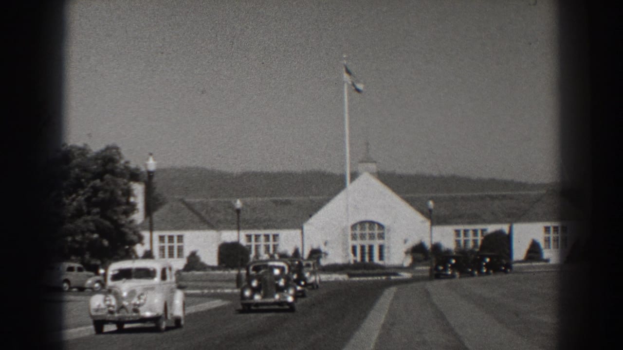 Period cars approach and pass a white gabled building with a flagpole in front.